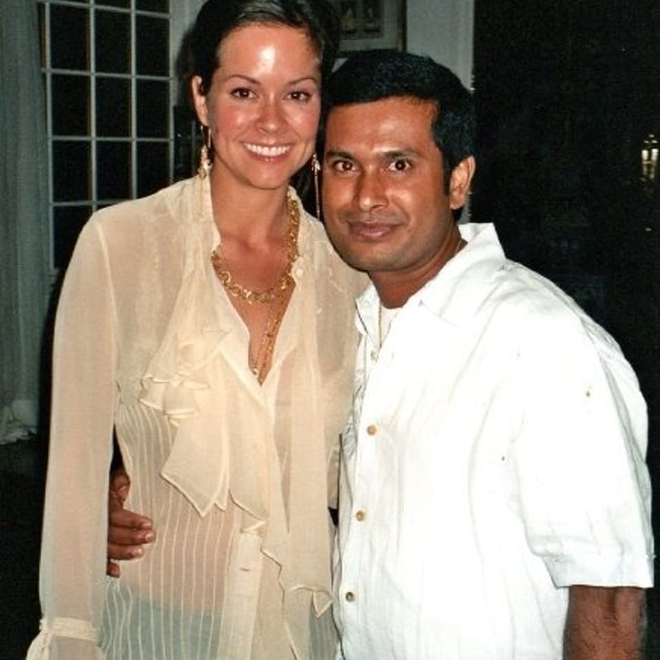 Smiling couple pose together indoors, with the woman wearing a sheer ruffled blouse and the man dressed in a white shirt.