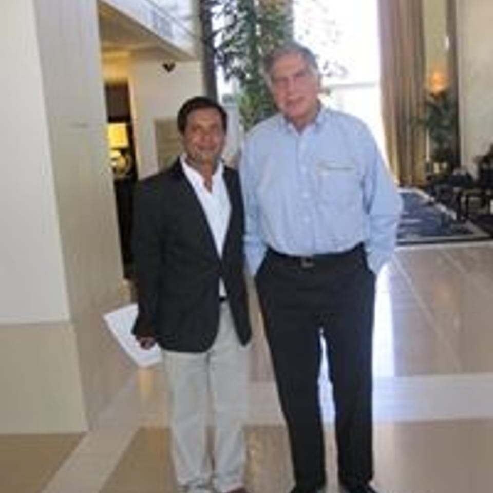 Two men stand together in a bright, upscale restaurant lobby, one wearing a dark blazer and the other a light blue shirt.