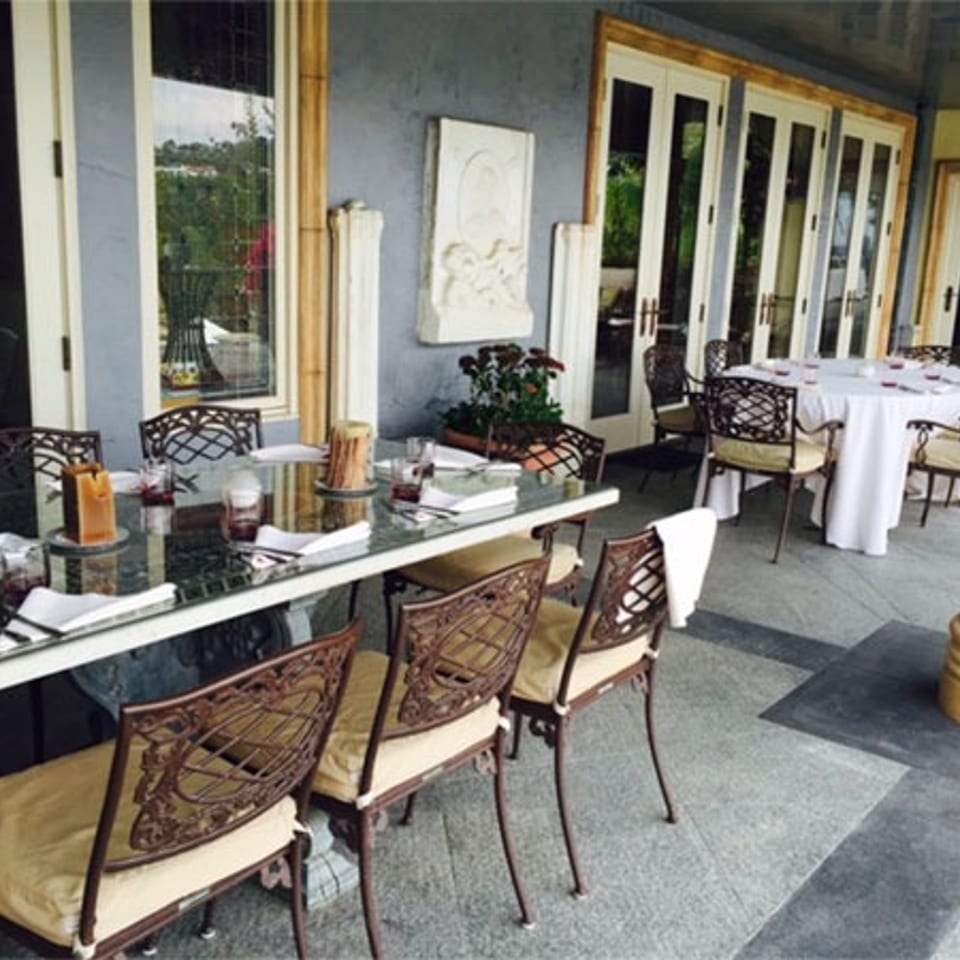 Elegant restaurant patio with set tables, wicker chairs, white linens, decorative columns, and greenery visible through glass doors.