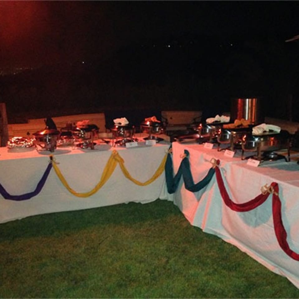 Outdoor buffet setup with chafing dishes and serving trays on draped tables decorated with colorful garlands.