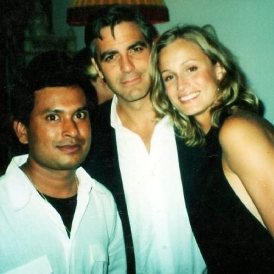Three smiling adults pose together indoors, with a man in a white shirt and a blonde woman embracing another man.