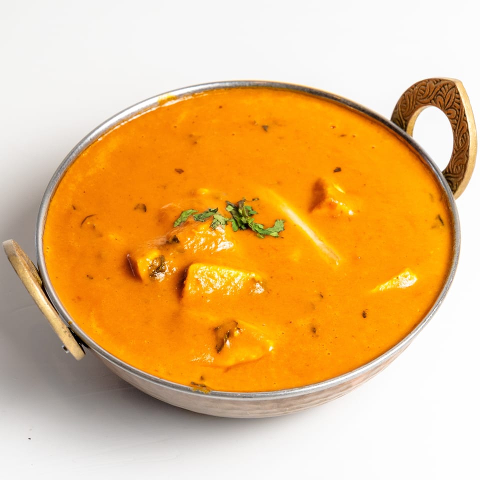 A metal bowl filled with orange curry, garnished with fresh green herbs, on a white background.
