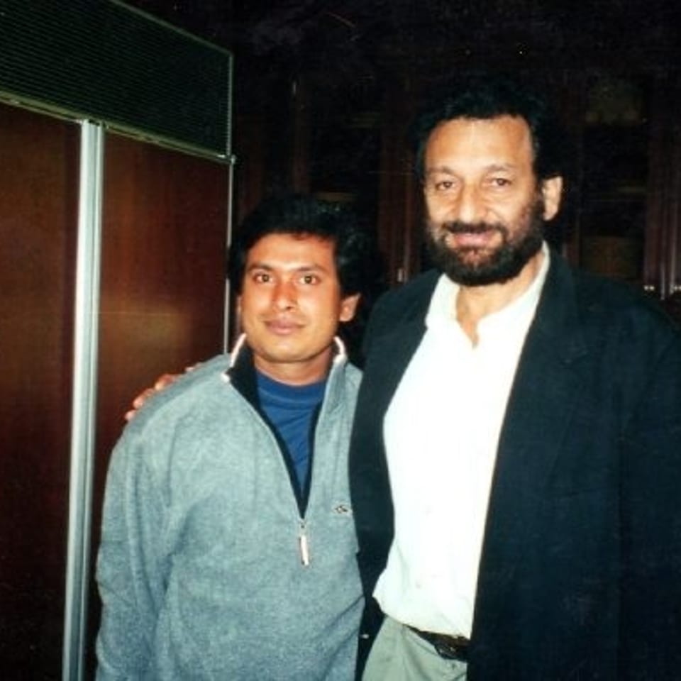 Two men pose together inside a dimly lit restaurant, with wood-paneled walls and a window in the background.
