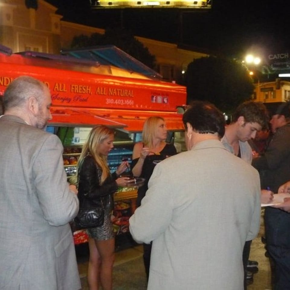 Crowd gathers around a bright red Indian food truck at night, with people ordering beneath illuminated palm trees.