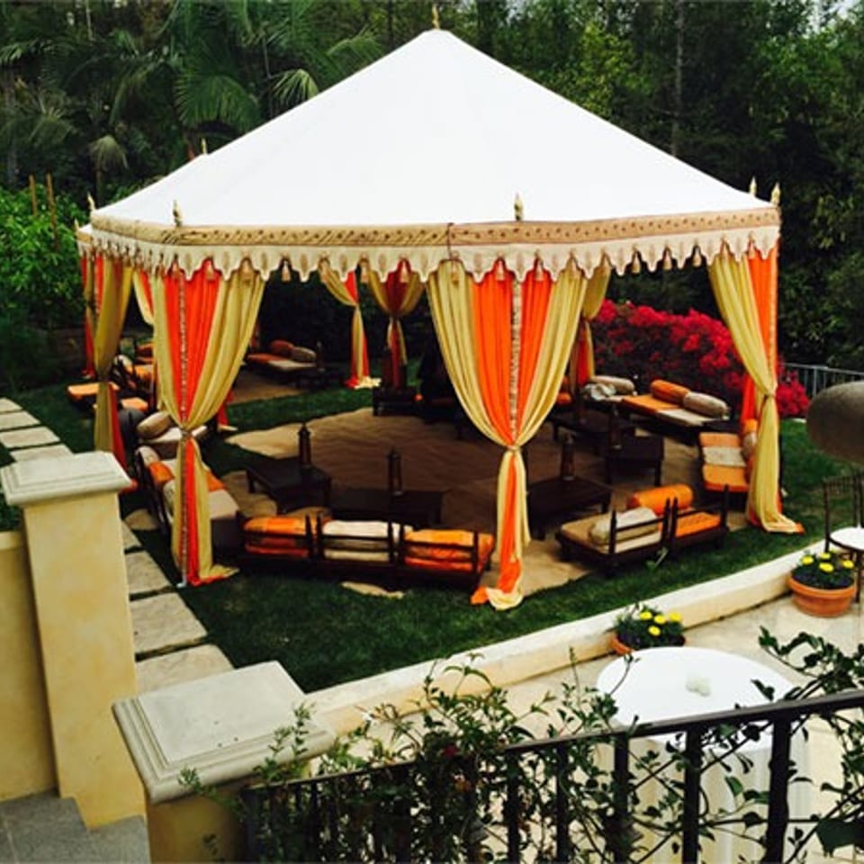 Elegant outdoor tent with orange and yellow drapes, lounge seating, and lush garden surroundings.