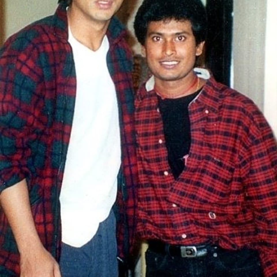 Two men stand indoors together, both wearing red plaid shirts, with one noticeably taller than the other.