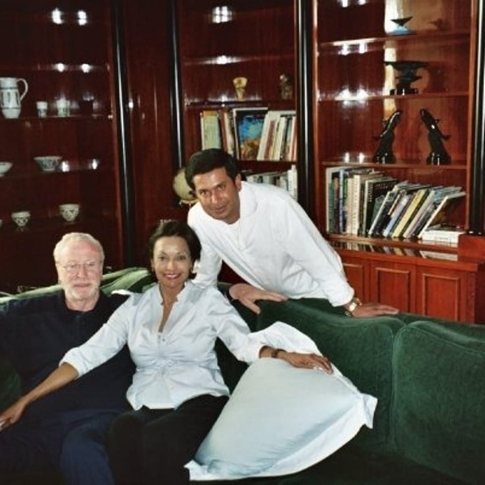 Three people relax on a green sofa in a warmly lit room with dark wood bookcases, books, ceramics, and decorative objects.