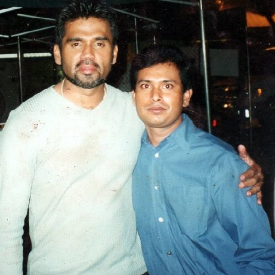 Two men pose together indoors, one wearing a light sweater and the other a blue button-down shirt.