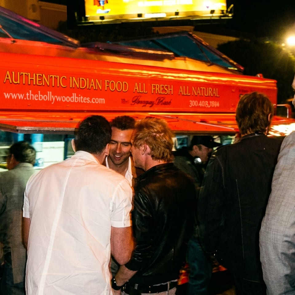 People gathered around a red food truck at night, serving "Authentic Indian Food" from Bollywood Bites.