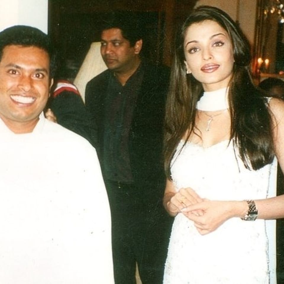 Three people pose indoors, including a smiling man in white and a woman wearing a white dress and scarf.