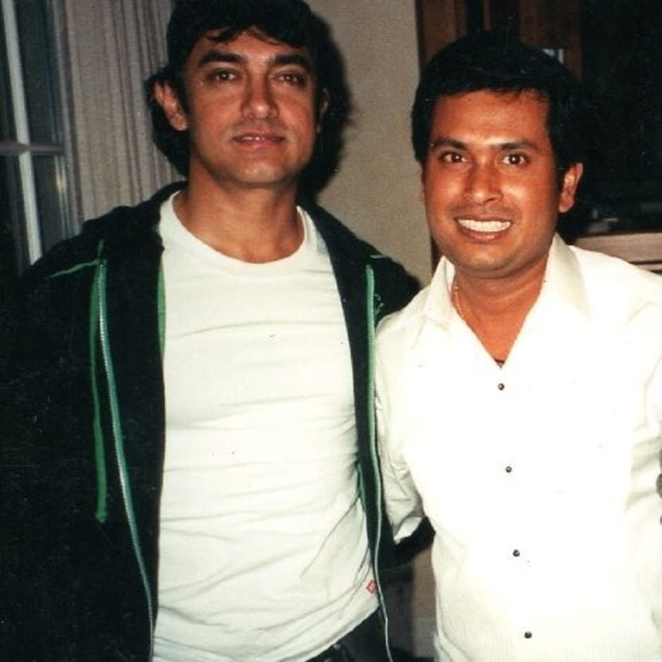 Two men pose together indoors, one wearing a black jacket and white shirt, the other dressed in a white button-down shirt.