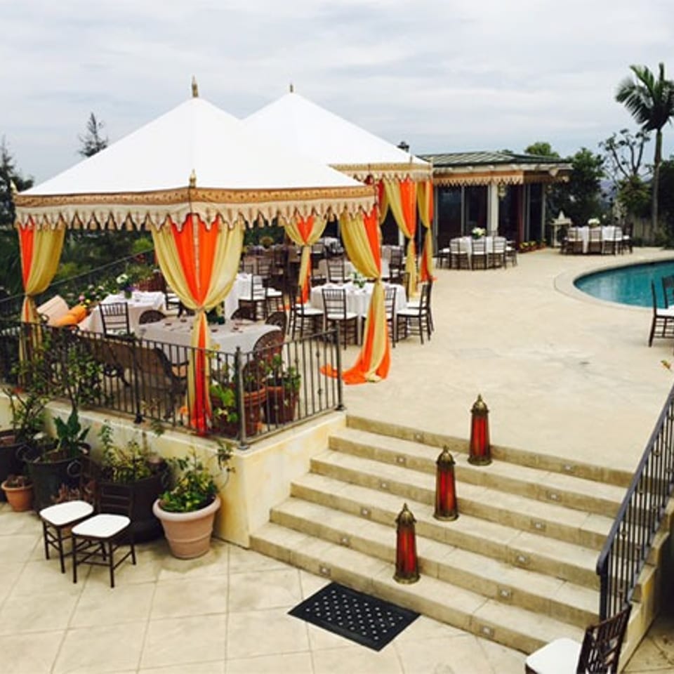 Outdoor restaurant terrace with colorful draped canopy, neatly set tables, pool, tropical plants, and stone steps.