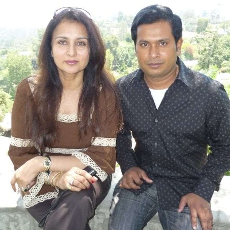 A woman and man pose together outdoors, seated before a scenic green landscape.