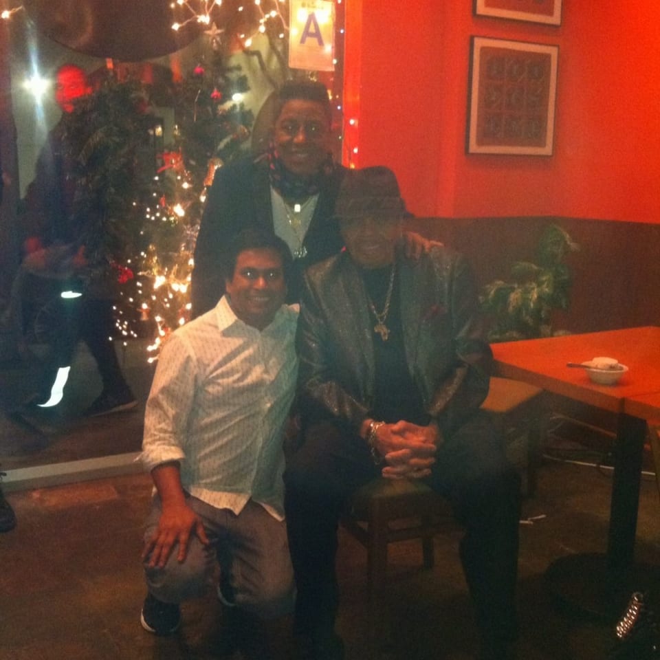 Three people pose inside a warmly lit restaurant beside a decorated Christmas tree, with string lights, framed artwork, and red walls.
