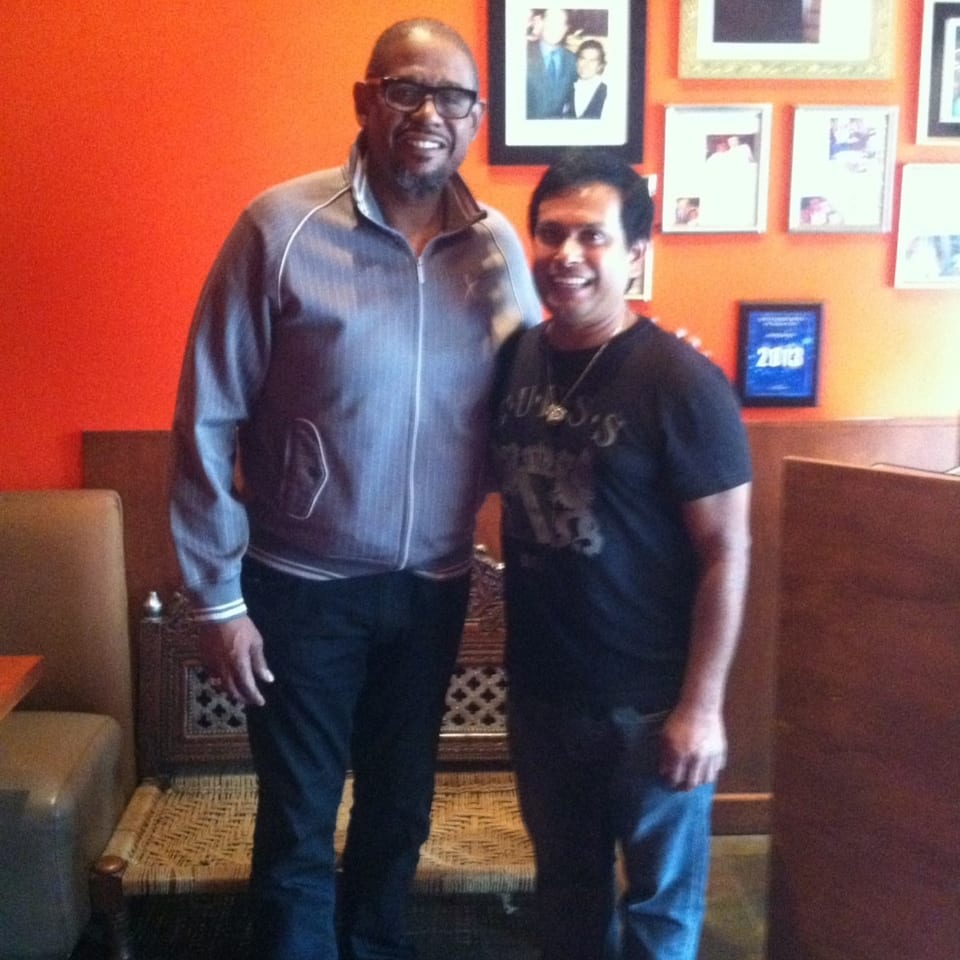 Two smiling men stand together in a restaurant lounge with orange walls, framed photographs, booths, and patterned carpeting.