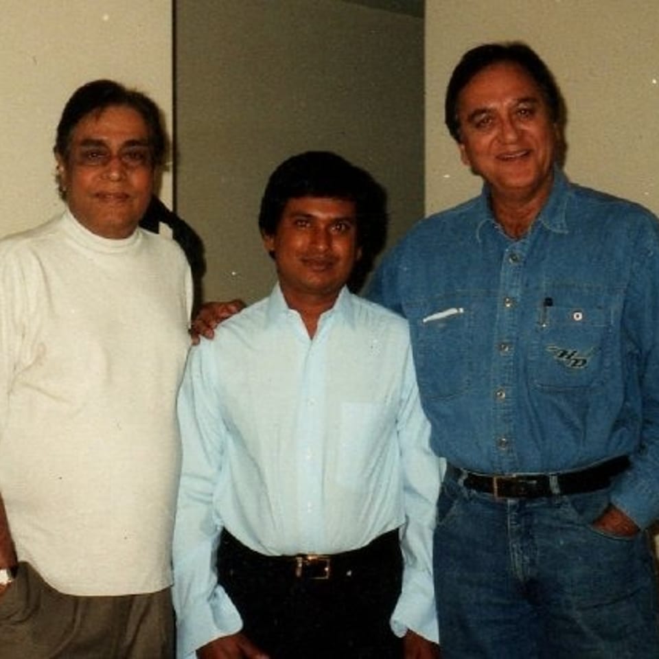 Three men pose indoors, wearing casual shirts and trousers against a neutral wall.