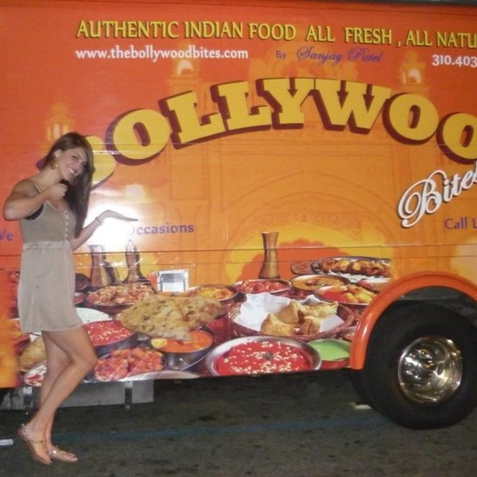 Woman posing beside a bright orange Bollywood Bites food truck displaying Indian dishes and catering information.