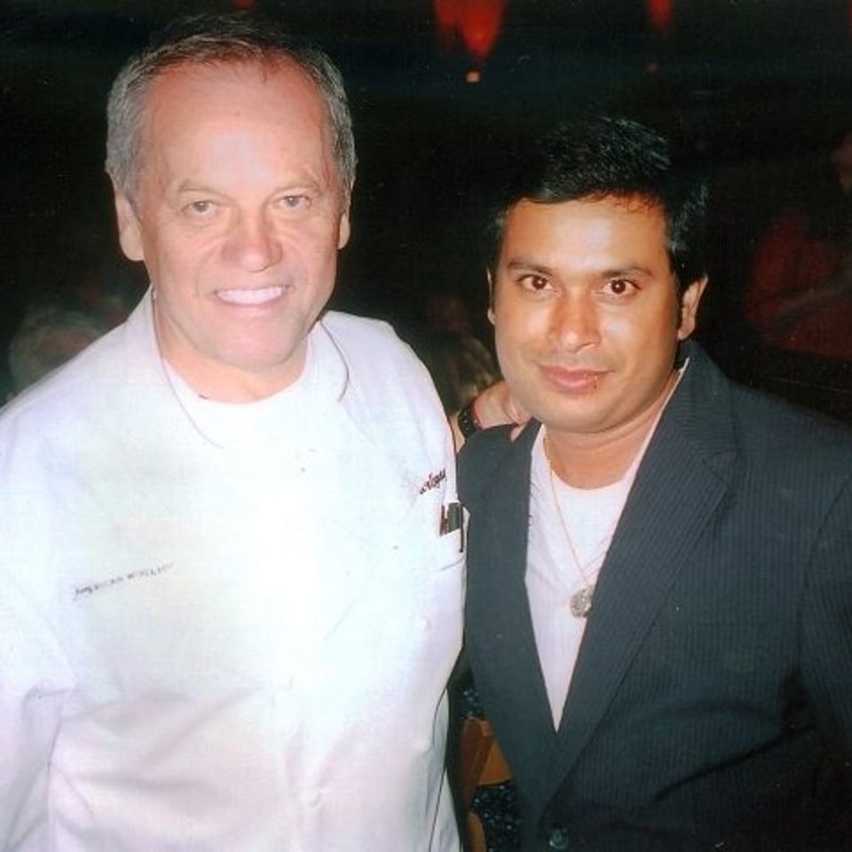 Two men pose together at a dimly lit restaurant event, one wearing a white chef’s jacket and the other a dark suit.