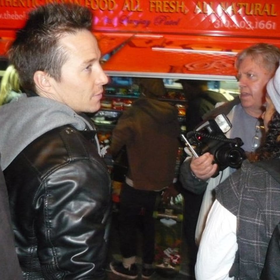 Man in a leather jacket speaks with a woman holding a video camera beside a red food truck.