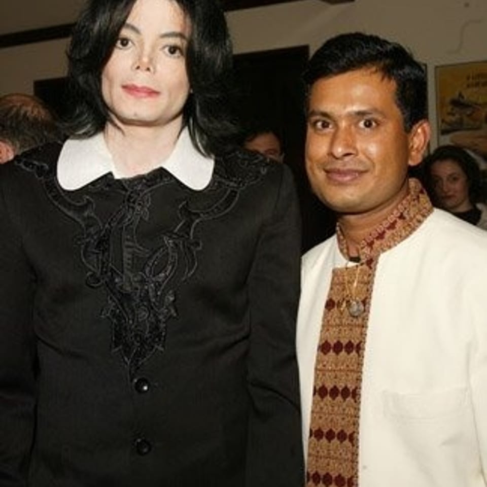 Michael Jackson and a man in traditional patterned attire pose together at a crowded indoor event.