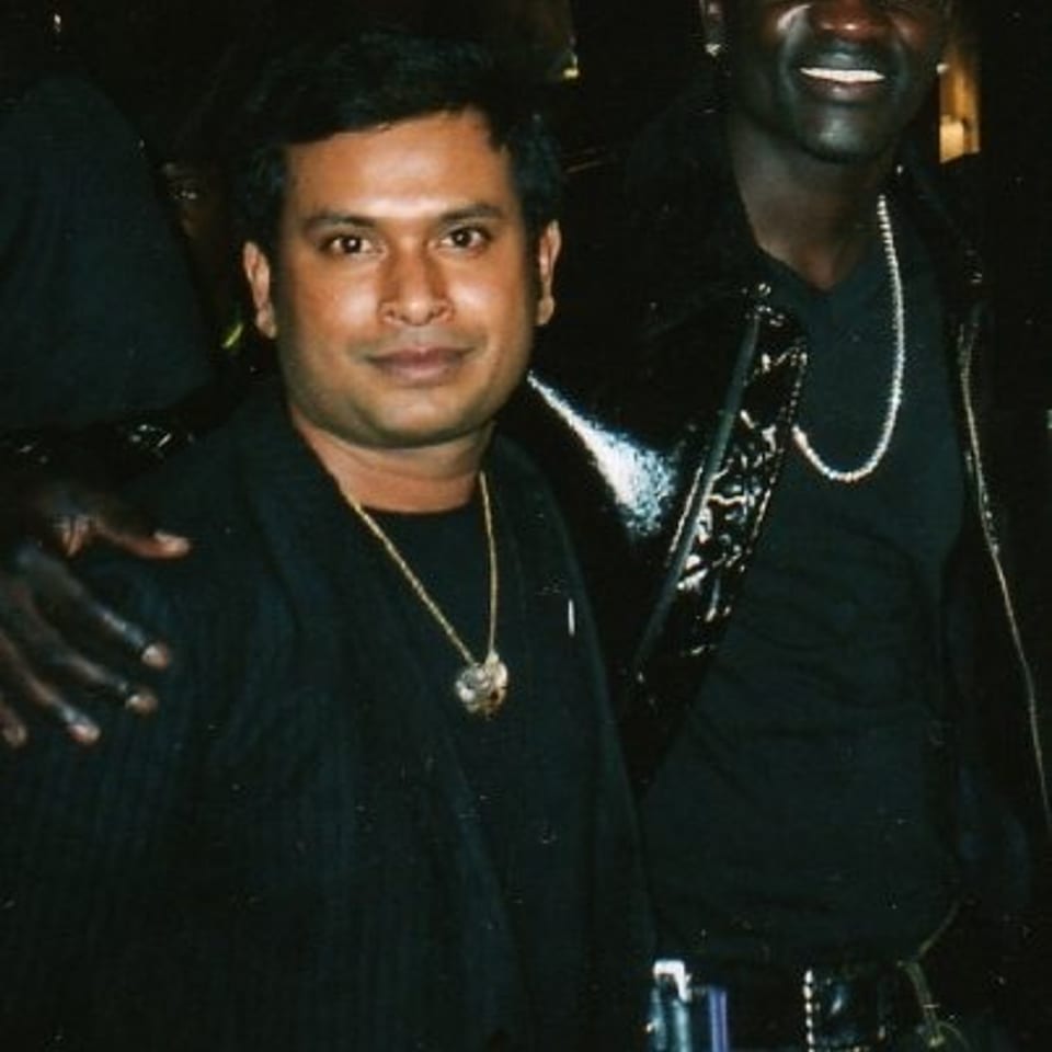 Two smiling men pose together in a dimly lit restaurant or nightlife setting, wearing dark clothing and necklaces.