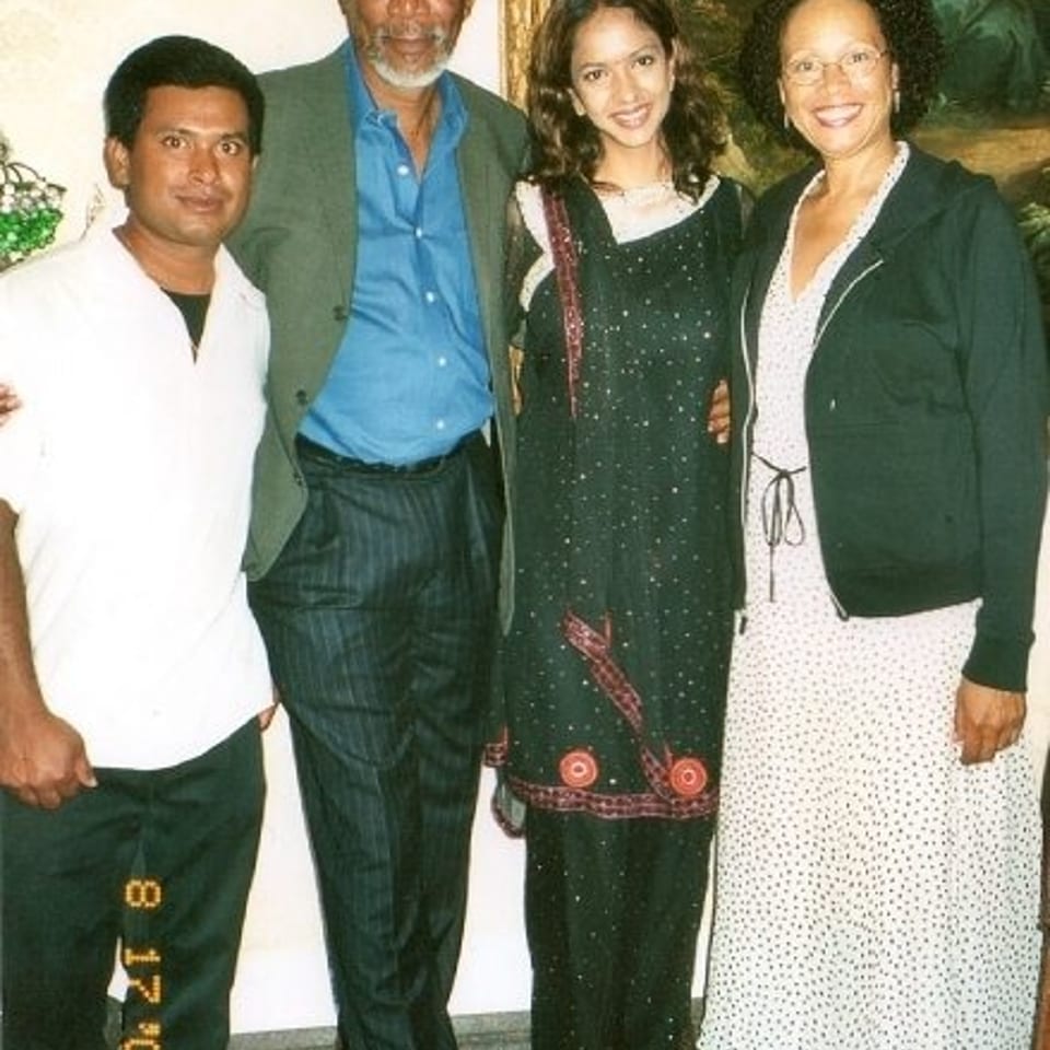 Four people pose together indoors beneath a large framed painting, with a woman wearing a traditional patterned dress.