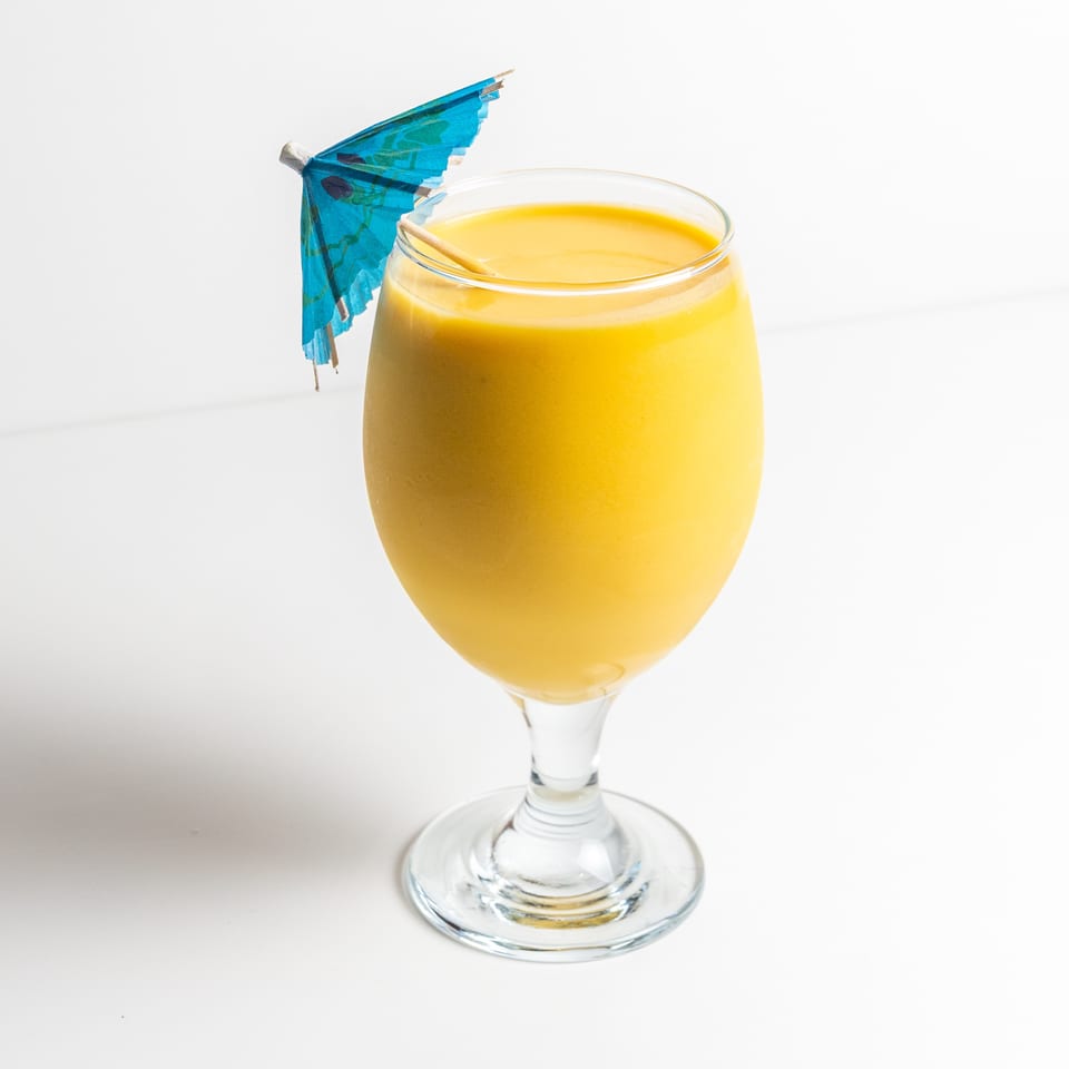 A vibrant yellow mango lassi in a stemmed glass, garnished with a small blue paper umbrella.