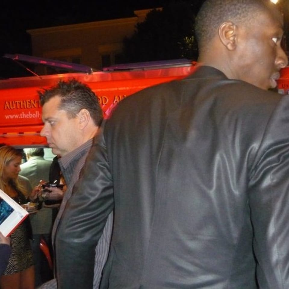Crowd gathers beside a brightly lit red double-decker bus at night, with people holding photographs and mingling.