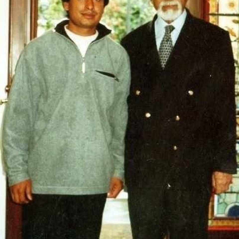 Two men stand indoors before a stained-glass window, one in casual clothing and the other in a formal suit.