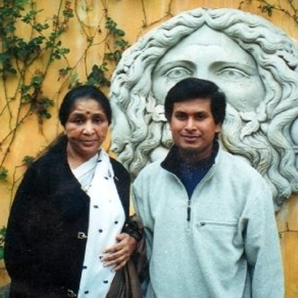 Two people pose beside a large sculpted classical face on an ivy-covered ochre wall.